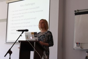 This picture shows a Caucasian woman (Josie Fraser) standing on the right hand side of the picture. She is standing at a lectern with a Macbook on it and a microphone pointing towards her. She has shoulder length blonde hair and is wearing a black, floral print dress. There are slides showing behind her. The text on the slides is: “Key challenges • I’ve never heard of open licences / OER •	 I don’t see the benefit of sharing resources openly / using OER •	 My resources are not good enough •	 My resources are too good •	 At the moment, I just do what I like and that’s working fine for me •	 Making, finding & accrediting = extra work”